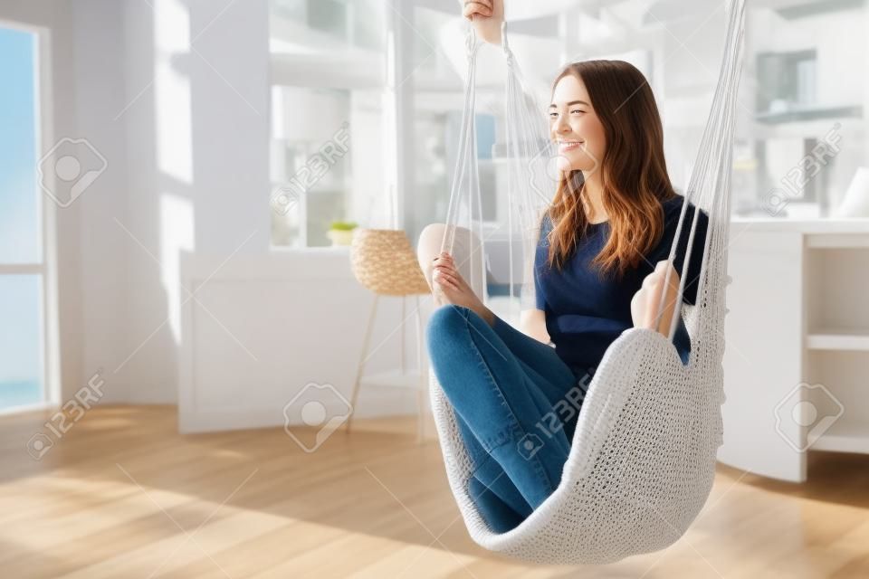 Young woman chilling at home in comfortable hanging chair in front of big window.