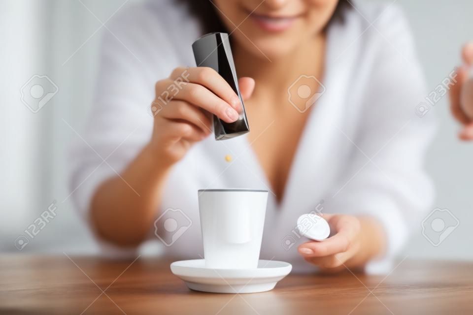 Close-up da mão da mulher jogando pílulas de sacarina no copo de café na cozinha em casa.