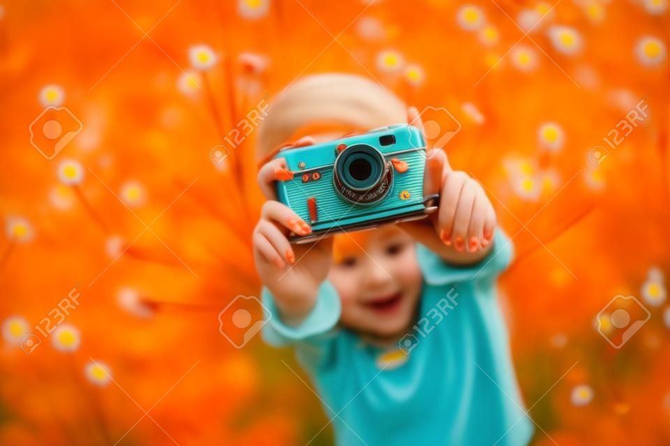 Młoda piękna dziewczyna w pomarańczowej koszulce trzymająca aparat retro w dłoniach leżąca na trawniku, gdzie rosną mlecze, kwitnące mlecze