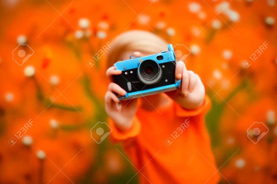Młoda piękna dziewczyna w pomarańczowej koszulce trzymająca aparat retro w dłoniach leżąca na trawniku, gdzie rosną mlecze, kwitnące mlecze