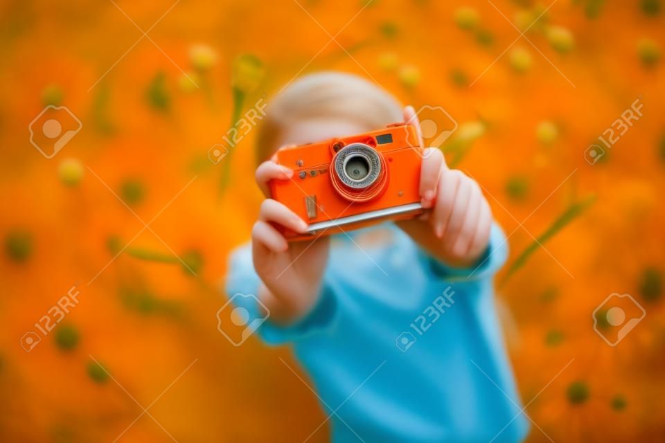 Young beautiful girl in an orange T-shirt holding retro camera in her hands lying on the lawn where dandelions grow, flowering dandelions