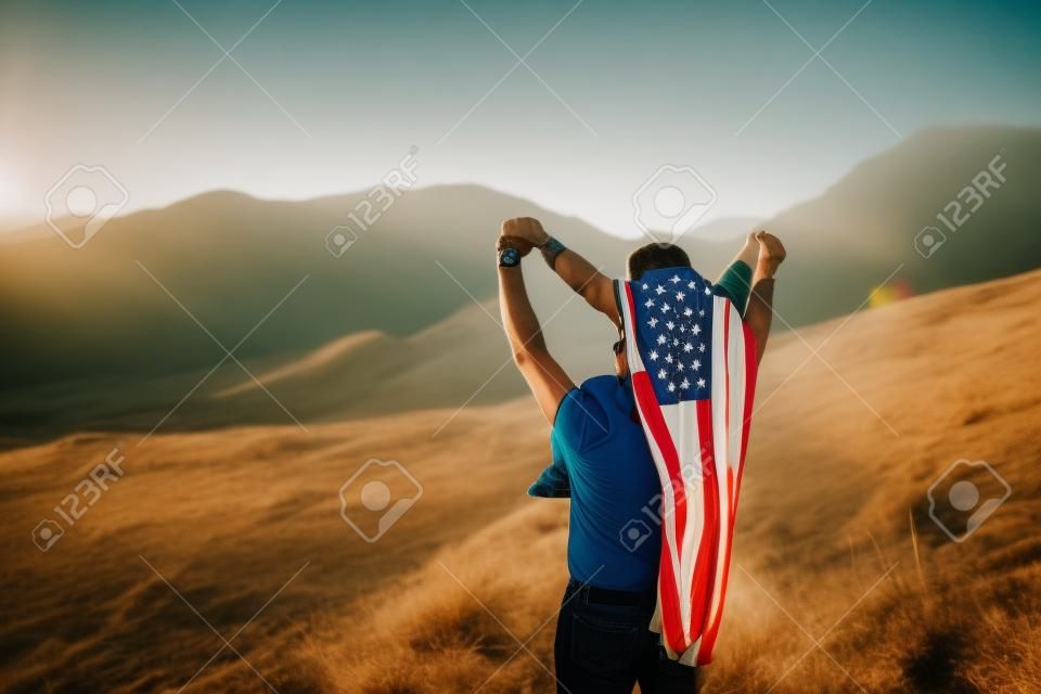 Man with child on his neck, boy on his back has an American flag on the background of nature.