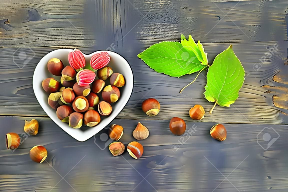 Fresh unshelled hazelnuts in a heart shaped dish with green leaves in an overhead flat lay still life on a wooden table with copyspace