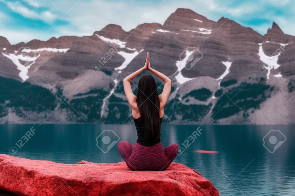 Jonge vrouw in donkerrode sportkleding yoga beoefenen in de natuur, zitten op de steen terug naar de camera en meditatie, bergmeer en bereik op de achtergrond, actief leven concept