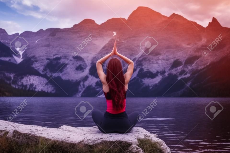 Jonge vrouw in donkerrode sportkleding yoga beoefenen in de natuur, zitten op de steen terug naar de camera en meditatie, bergmeer en bereik op de achtergrond, actief leven concept