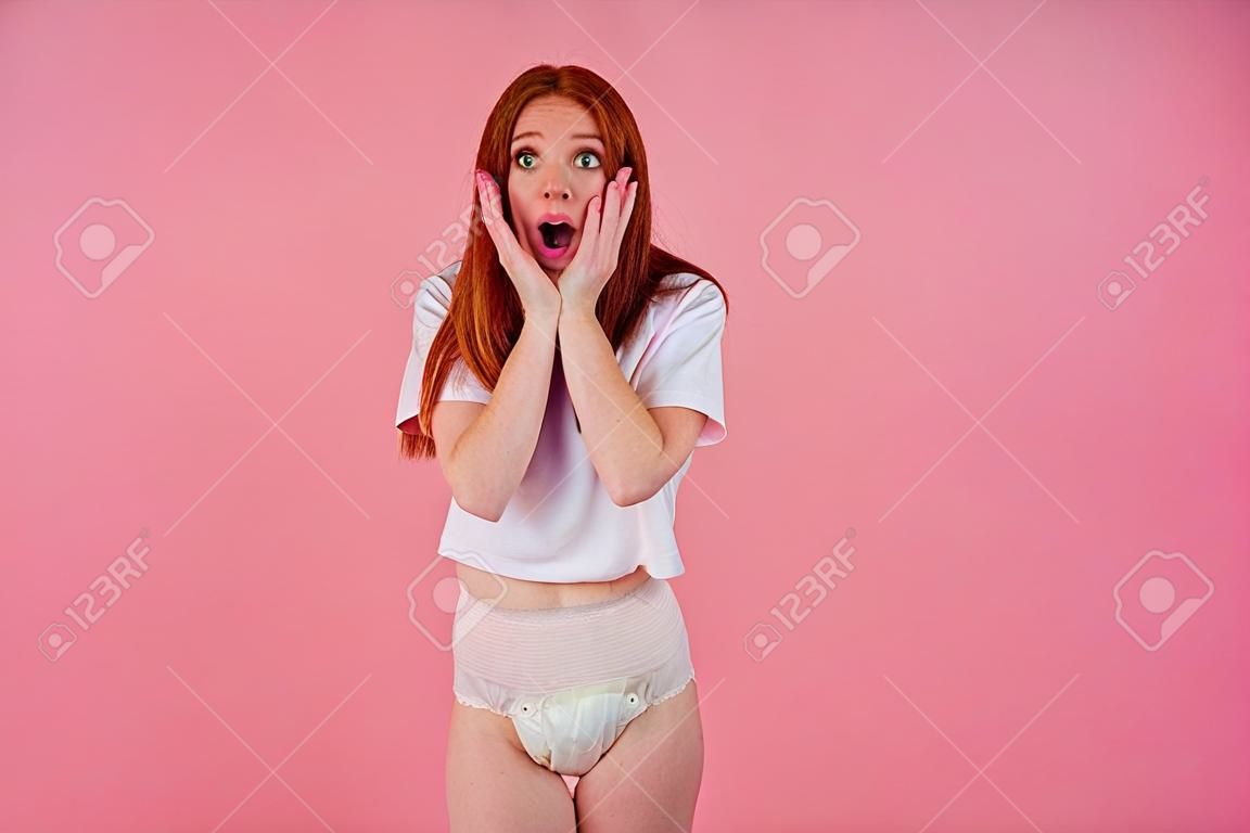 스튜디오 분홍색 배경에서 요실금 기저귀를 입은 젊은 놀랍고 놀란 빨간 머리 생강 여자. 수치심과 당혹감을 느끼는