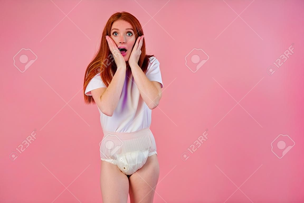 młoda zdumiona i zaskoczona rudowłosa ruda kobieta nosząca pieluchę dla nietrzymania moczu w studio różowym tle. uczucie wstydu i zakłopotania