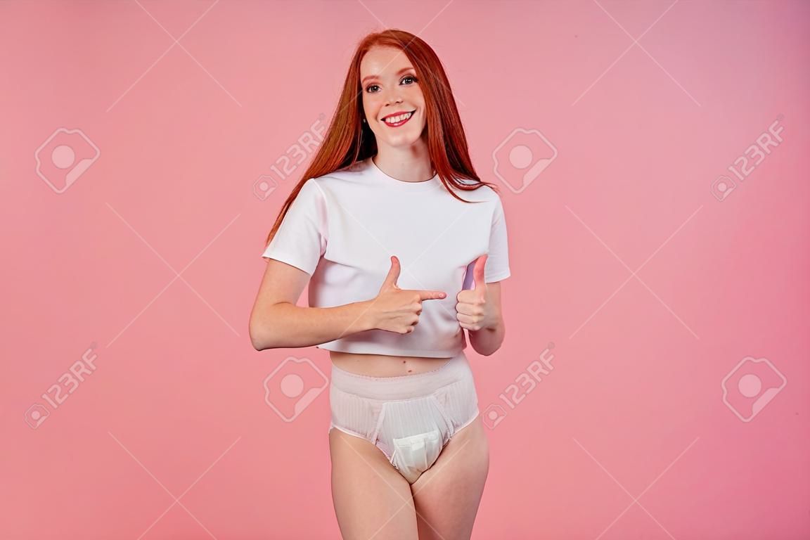 glückliche junge rothaarige Ingwerfrau, die Inkontinenzwindel im Studio rosa Hintergrund trägt, zeigt Daumen hoch