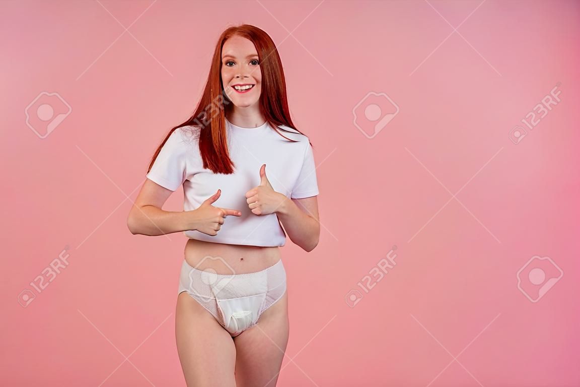 スタジオピンクの背景ショーイム親指で失禁のおむつを身に着けている幸せな若い赤毛の生姜の女性