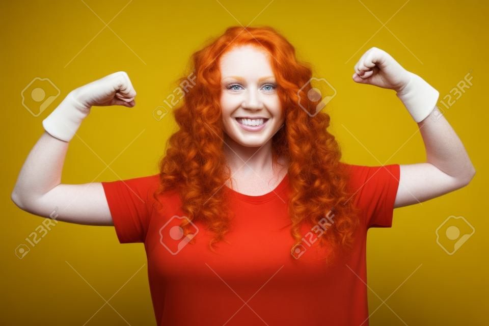 donna di zenzero riccia dai capelli rossi che mostra i muscoli delle mani di allenamento nella parete di fondo gialla dello studio