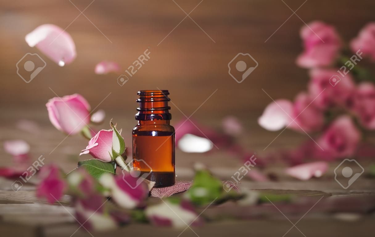 Closeup de garrafa com óleo essencial de rosa com folhas caindo no fundo de madeira.