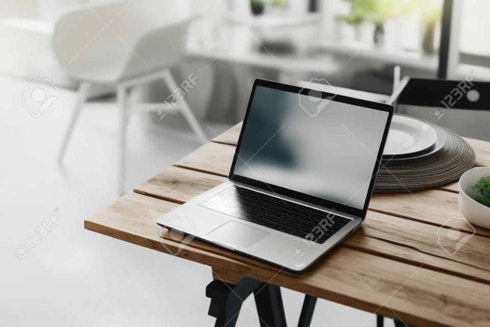Laptop mit weißem Bildschirm auf dem rustikalen Holztisch im Küchenraum, mit verschwommenem Interieur im Hintergrund, Attrappe. Foto in hoher Qualität
