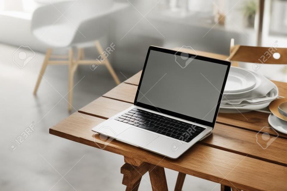 キッチンルームの素朴な木製テーブルに白い画面があり、背景にぼやけたインテリアがあるノートパソコンがモックアップされています。高品質の写真