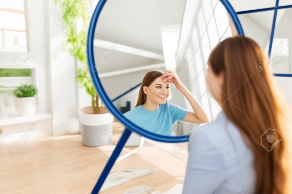 Attraente giovane donna caucasica in cuffia che tocca il viso e guarda lo specchio in soggiorno a casa in una giornata di sole. Concetto di bellezza e cura della pelle. Foto di alta qualità