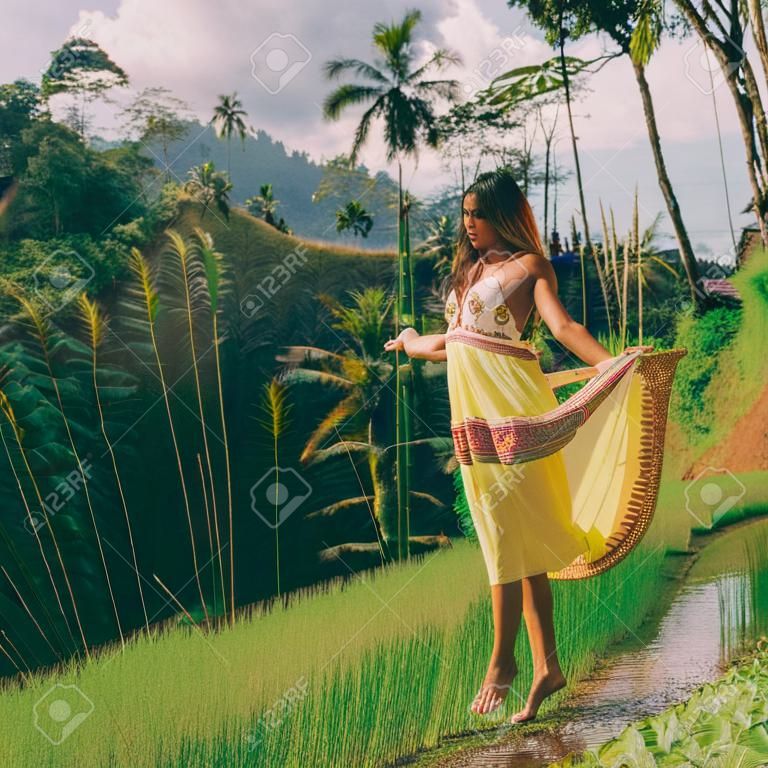 Beautiful young woman in yellow fashion dress. Girl travel and explore world. Typical Asian hillside with rice farming, mountain shape green cascade rice field terraces paddies. Ubud, Bali, Indonesia.