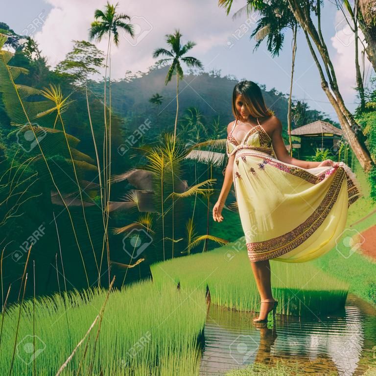 Beautiful young woman in yellow fashion dress. Girl travel and explore world. Typical Asian hillside with rice farming, mountain shape green cascade rice field terraces paddies. Ubud, Bali, Indonesia.