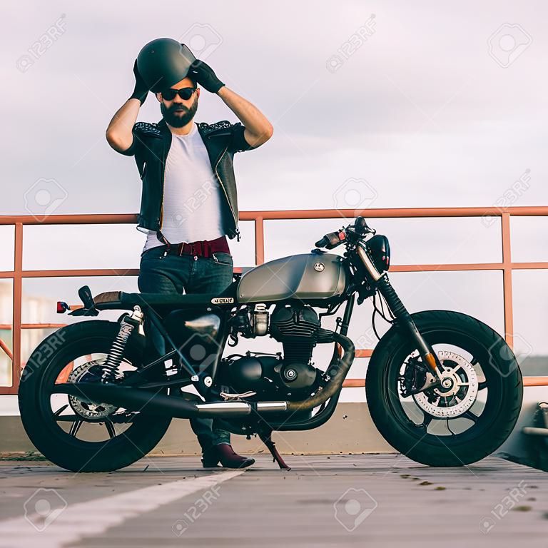 Handsome happy rider guy with beard and mustache put on black moto helmet and sit on his classic style biker cafe racer motorcycle. Bike custom made in vintage garage. Brutal fun urban lifestyle.