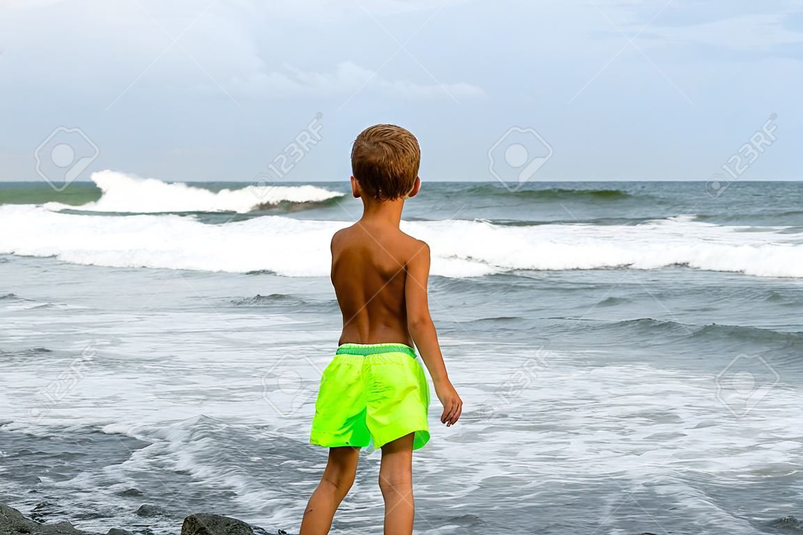 002_ Una veduta di spalle di un ragazzo in costume da bagno, che sta in piedi sulla spiaggia e guarda il mare durante un temporale