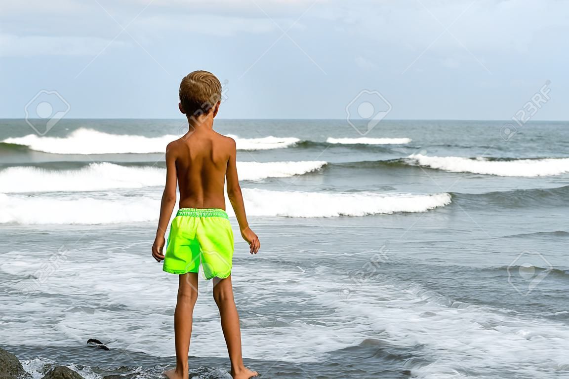 002_ Una veduta di spalle di un ragazzo in costume da bagno, che sta in piedi sulla spiaggia e guarda il mare durante un temporale