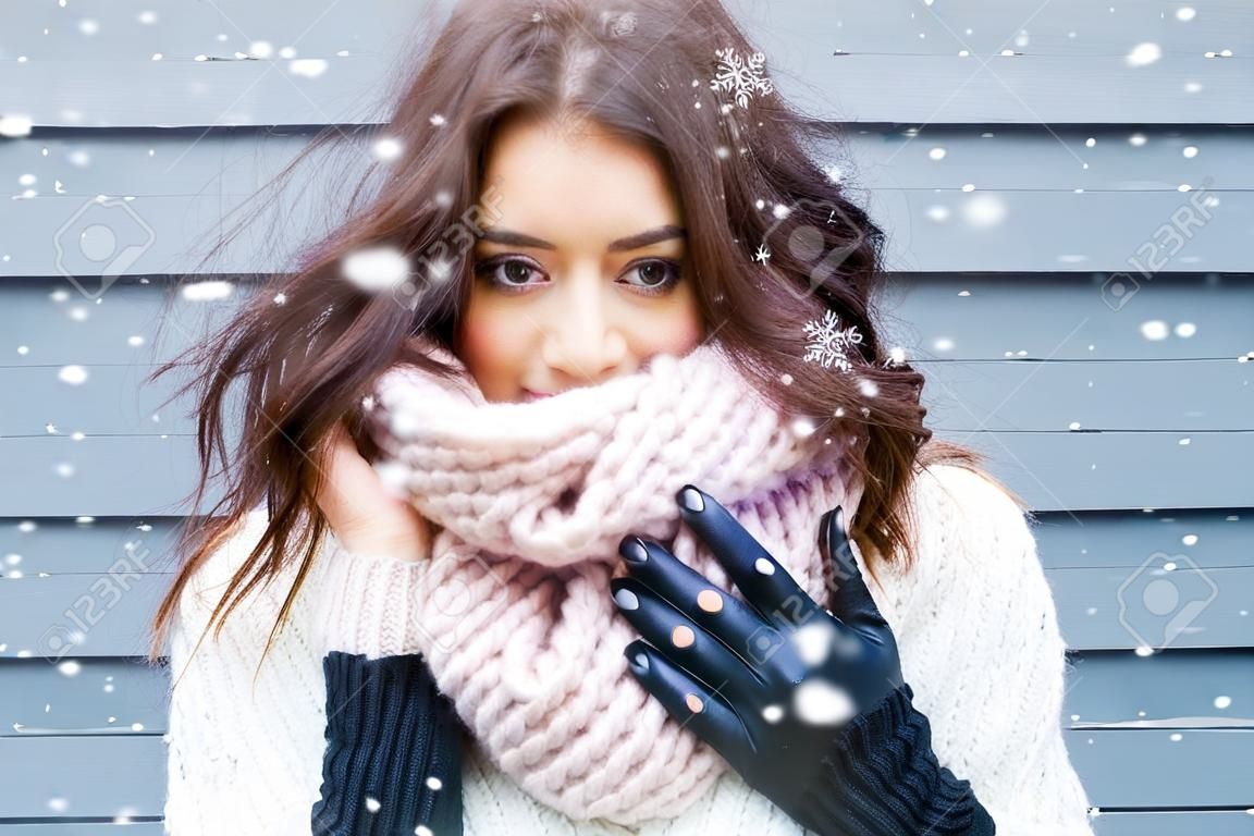 身に着けている若い美しいブルネットの女性の冬の肖像ニット スヌードが雪で覆われています。雪が降る冬美容ファッションのコンセプトです。