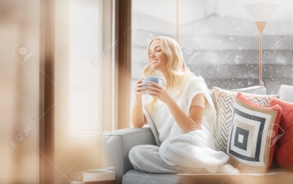 Junge schöne blonde frau mit einer tasse kaffee durch das fenster zu hause sitzen im wohnzimmer. winterschneelandschaft blick. lazy day off-konzept