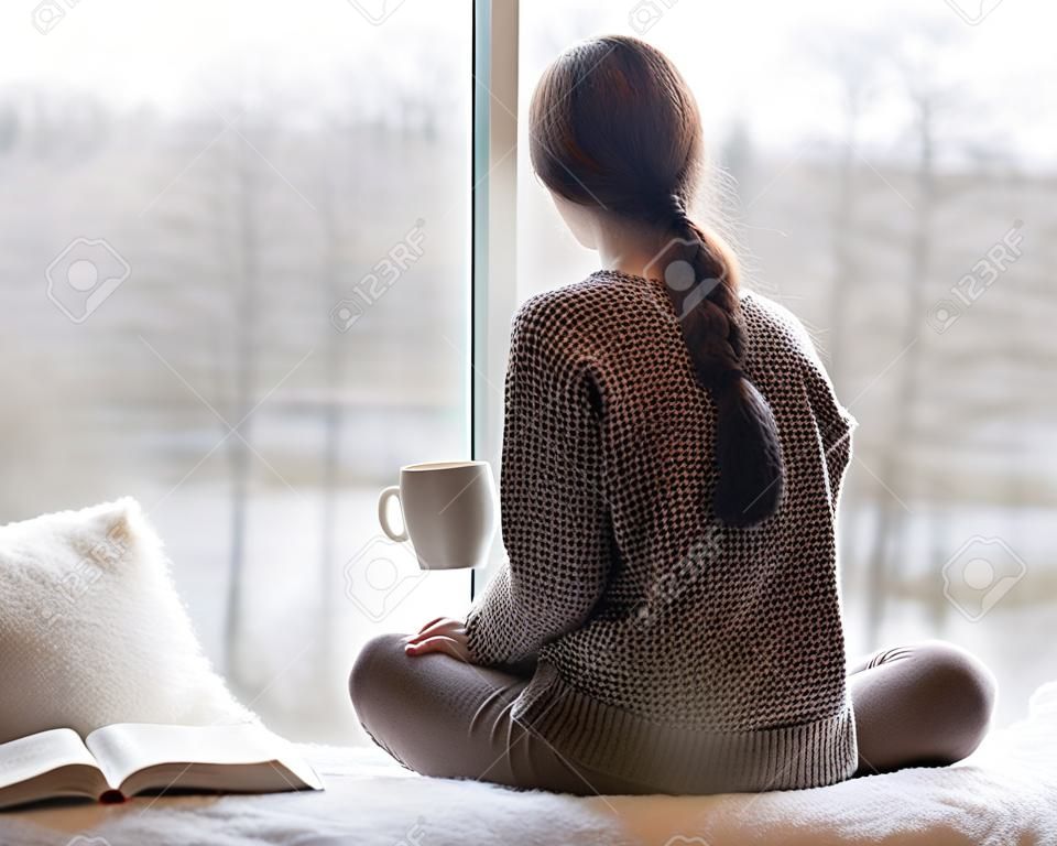 Thoughtful young brunette woman with book and cup of coffee looking through the window, blurry winter forrest landscape outside