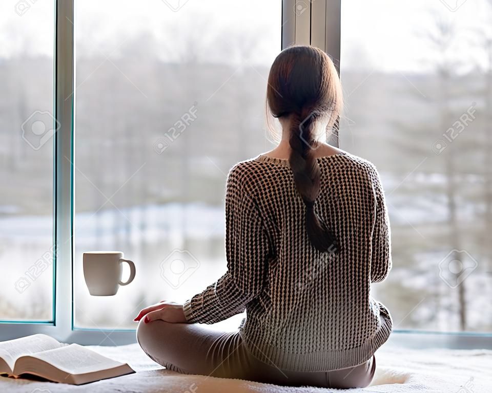 Задумчивый молодая брюнетка женщина с книгой и чашкой кофе, глядя через окно, размытый зимний пейзаж forrest вне