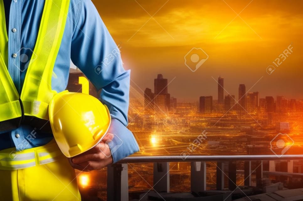 Engineer holding a yellow helmet with a backdrop of the city and industry.