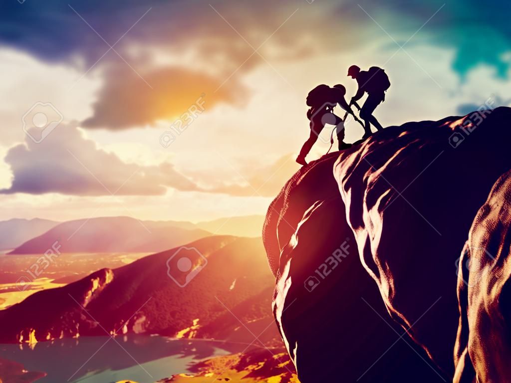Hikers climbing on rock, mountain at sunset, one of them giving hand and helping to climb  Help, support, assistance in a dangerous situation