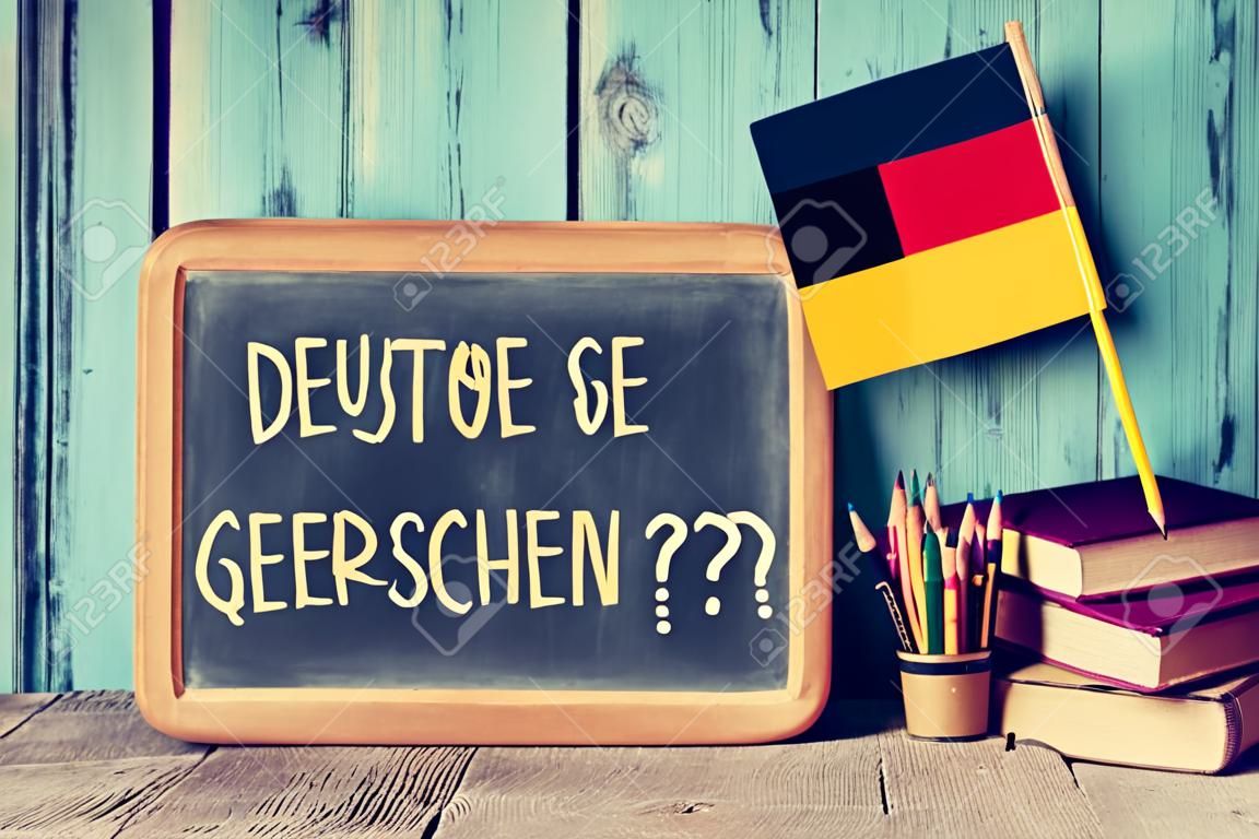 um quadro-negro com a pergunta sprechen sie deutsch? você fala alemão? escrito em alemão, um pote com lápis, alguns livros e a bandeira da Alemanha, em uma mesa de madeira