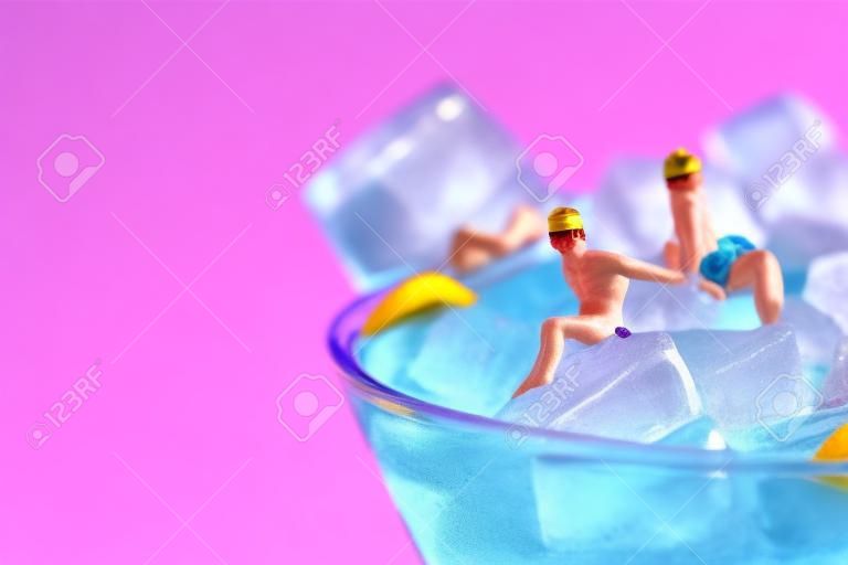 enkele miniatuur mannen in badpak ontspannend op de ijsblokjes van een blauwe cocktail geserveerd in een cocktail glas, tegen een roze achtergrond met wat lege ruimte aan de linkerkant