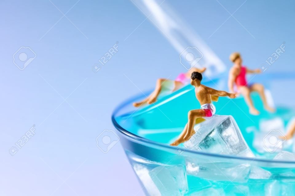 some miniature men wearing swimsuit relaxing on the ice cubes of a blue cocktail served in a cocktail glass, against a pink background with some blank space on the left