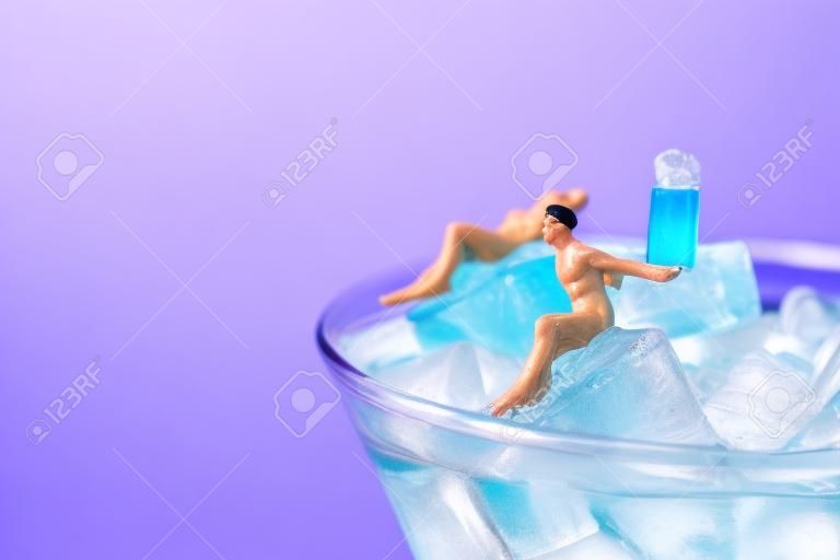 some miniature men wearing swimsuit relaxing on the ice cubes of a blue cocktail served in a cocktail glass, against a pink background with some blank space on the left