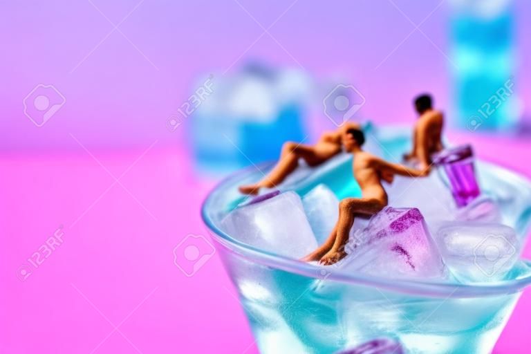 Einige miniaturmänner tragen einen badeanzug, der sich auf den eiswürfeln eines blauen cocktails entspannt, der in einem cocktailglas vor einem rosa hintergrund mit einer leerstelle auf der linken seite serviert wird