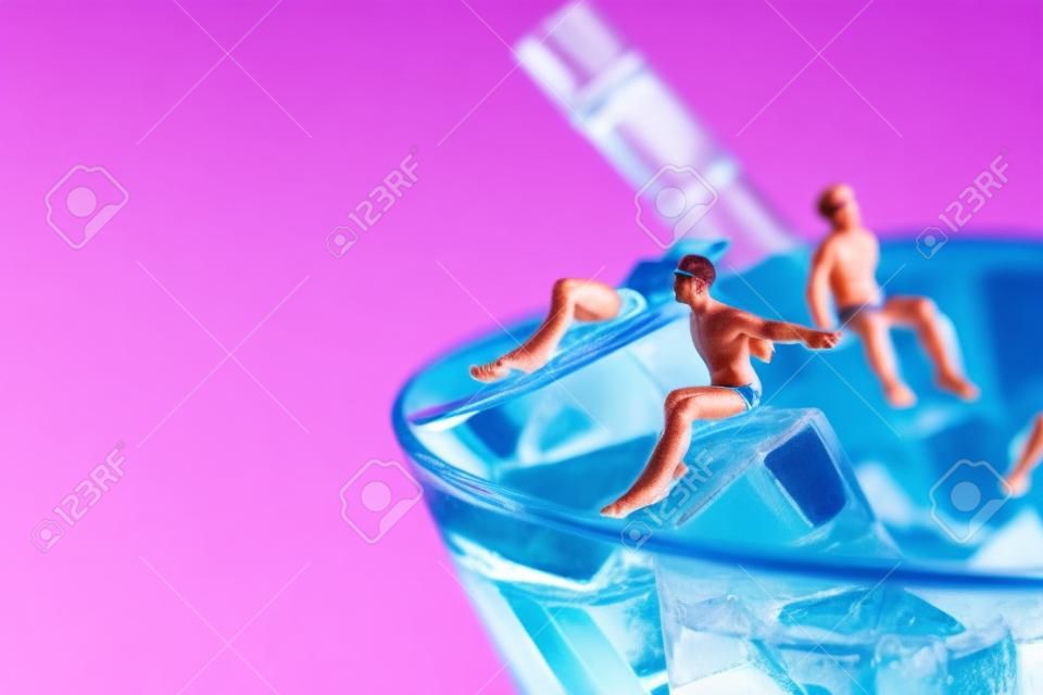 néhány fürdőruhát viselő miniatűr férfi egy kék koktél jégkockáin pihentető koktélpohárban, rózsaszín háttér előtt, bal oldalon üres hely
