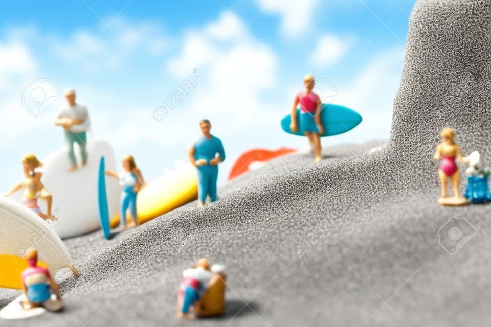 alcune persone in miniatura diverse che indossano il costume da bagno che trasportano tavole da surf o si rilassano sulla sabbia della spiaggia accanto a una conchiglia