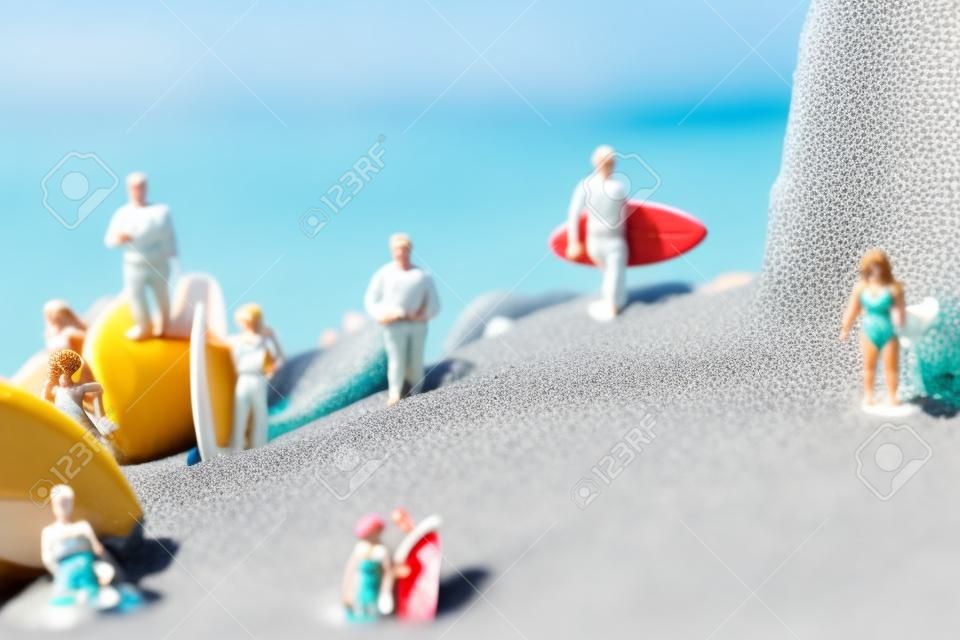 alcune persone in miniatura diverse che indossano il costume da bagno che trasportano tavole da surf o si rilassano sulla sabbia della spiaggia accanto a una conchiglia
