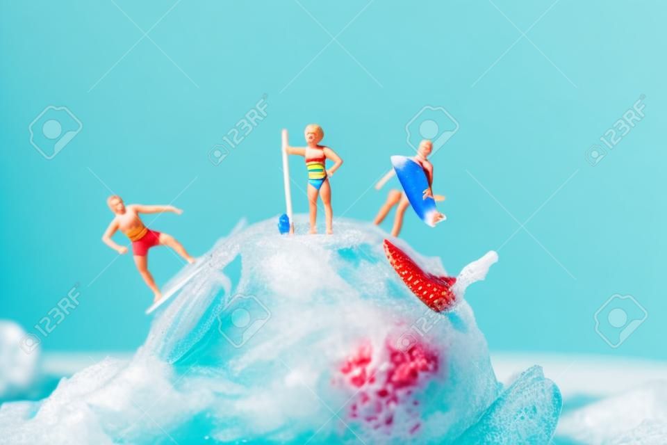 closeup of some miniature people in swimsuit surfing on a strawberry ice cream ball, against a blue background with some blank space