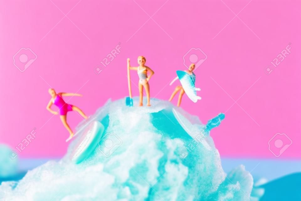 closeup de algumas pessoas em miniatura em maiô surfando em uma bola de sorvete de morango, contra um fundo azul com algum espaço em branco