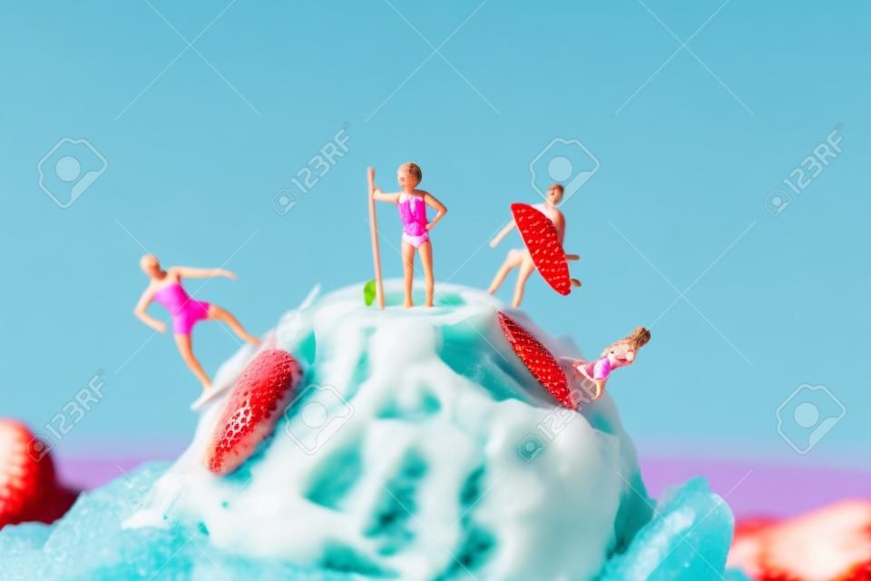 closeup of some miniature people in swimsuit surfing on a strawberry ice cream ball, against a blue background with some blank space