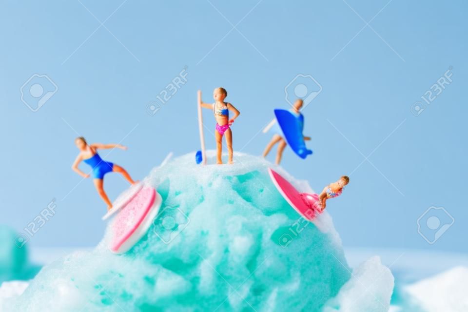 いくつかの空白を持つ青い背景に対して、イチゴアイスクリームボールでサーフィン水着でいくつかのミニチュアの人々のクローズアップ