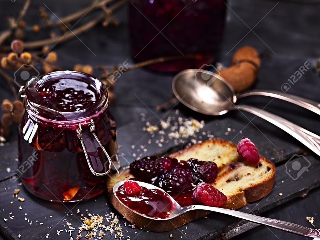 glass jar with raspberry jam and a slice of bread with a spoon on a black table