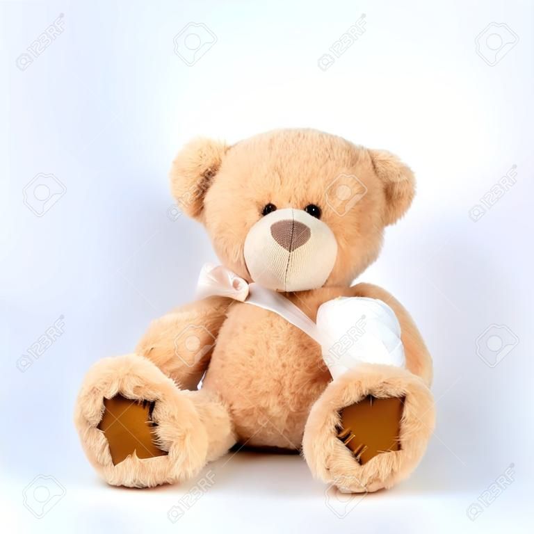 large beige teddy bear with patches sits on a white background, left paw is bandaged with a white medical bandage, concept of pediatrics, treatment of animals