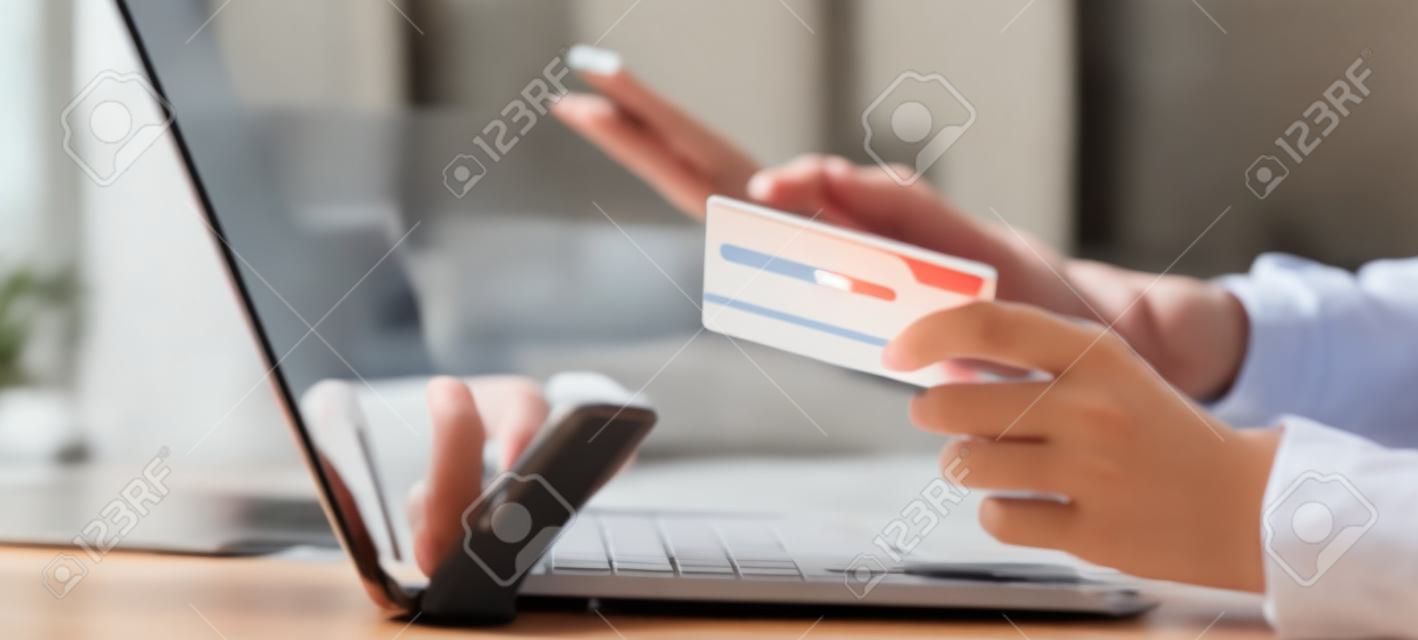 Closeup young asian business woman using smart phone and holding credit card while online shopping and payment with laptop computer on desk at home, female holding debit card, communication concept.