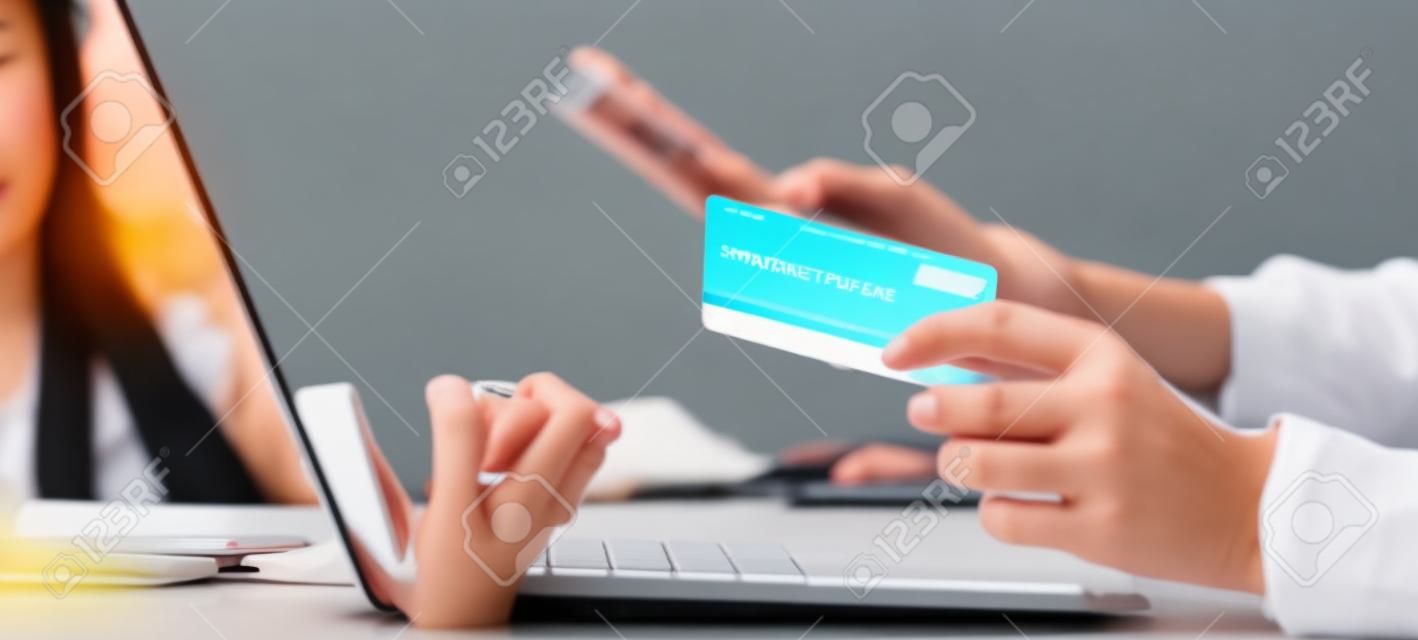 Closeup young asian business woman using smart phone and holding credit card while online shopping and payment with laptop computer on desk at home, female holding debit card, communication concept.