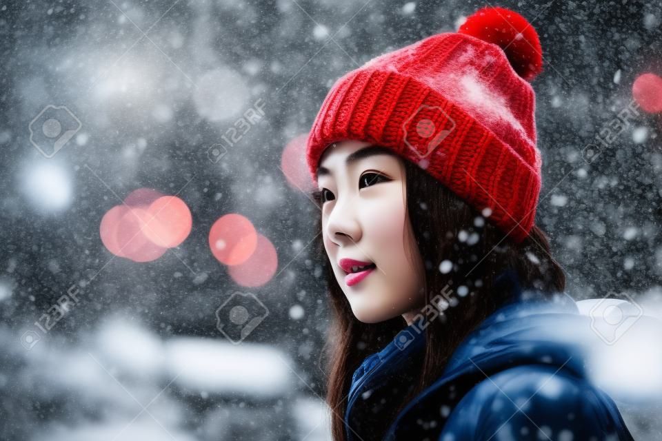 young beautiful and happy Asian Korean woman in winter hat and coat walking at city street under heavy snowfall in holidays travel and fashion concept