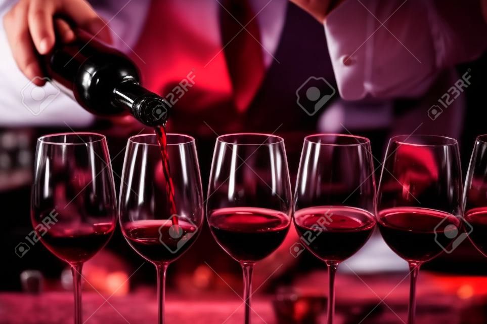 Bartender derrama vinho tinto em copos no bar.Sommelier masculino derramando vinho tinto em taças de vinho de haste longa.