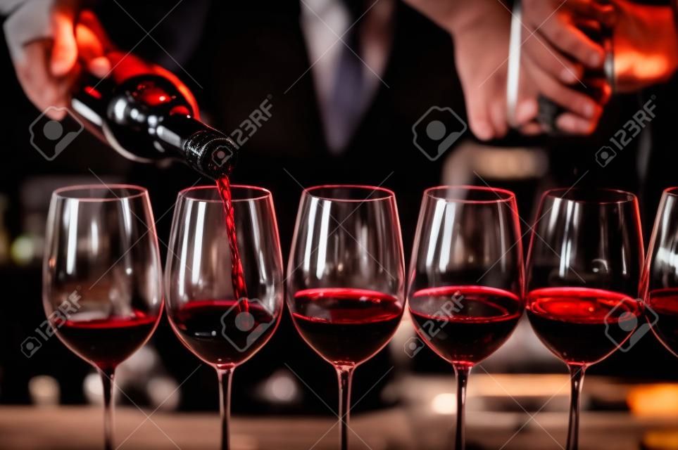 Bartender derrama vinho tinto em copos no bar.Sommelier masculino derramando vinho tinto em taças de vinho de haste longa.