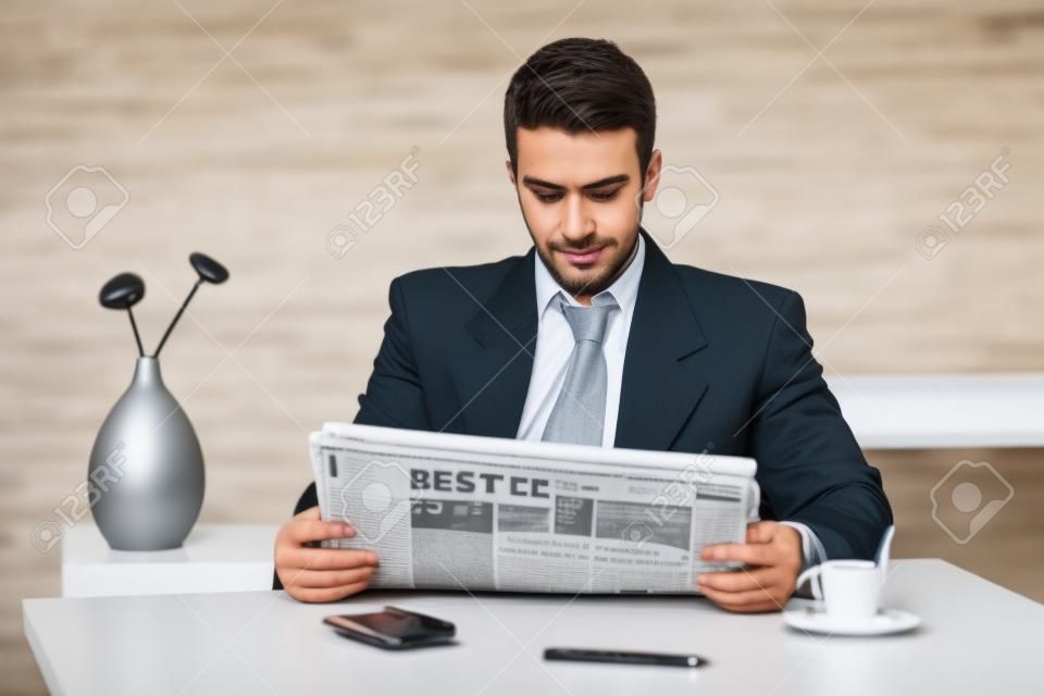 Young businessman having coffee break, sitting at office desk and reading newspaper.
