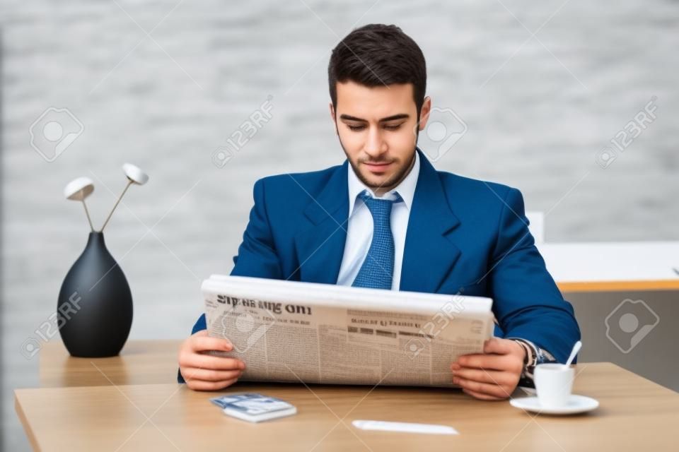 Young businessman having coffee break, sitting at office desk and reading newspaper.
